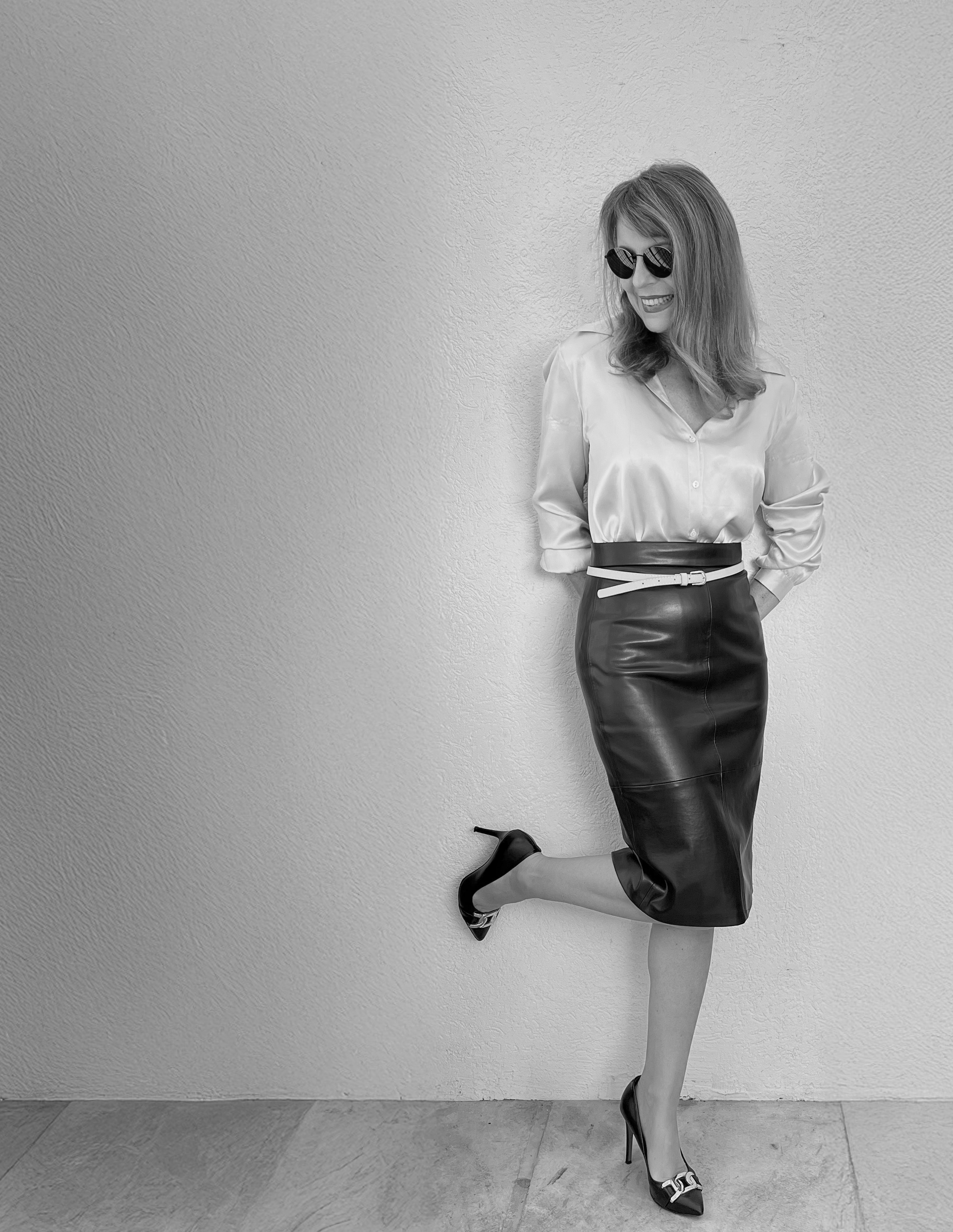 Anna Tendera in a black-and-white portrait, leaning against a wall with sunglasses and a playful kick